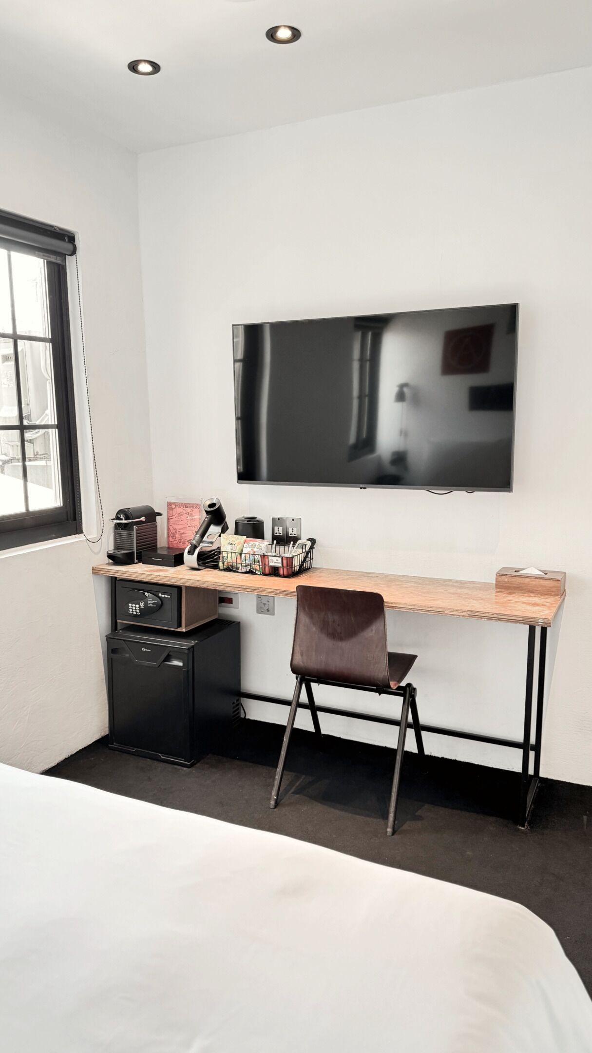 A modern hotel room with a large bed, a desk with a chair, a television, a mini-fridge, and a window with blinds.