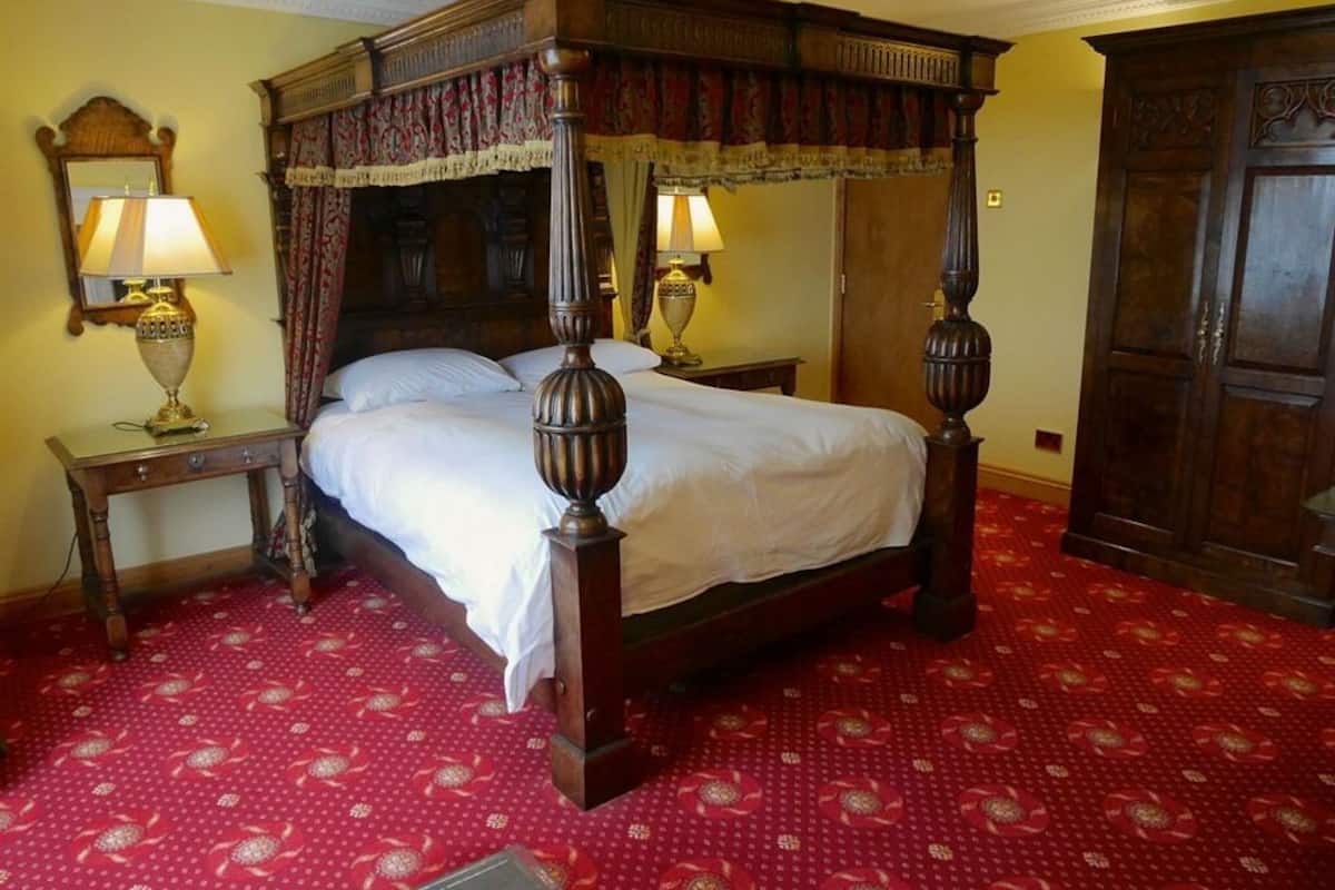 A four-poster bed with a wooden frame and white bedding, two bedside tables with lamps, and a red patterned carpet.