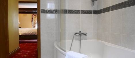 Un baño de hotel con ducha de mampara de vidrio, una bañera blanca y un pasillo con alfombra roja que conduce a un dormitorio.