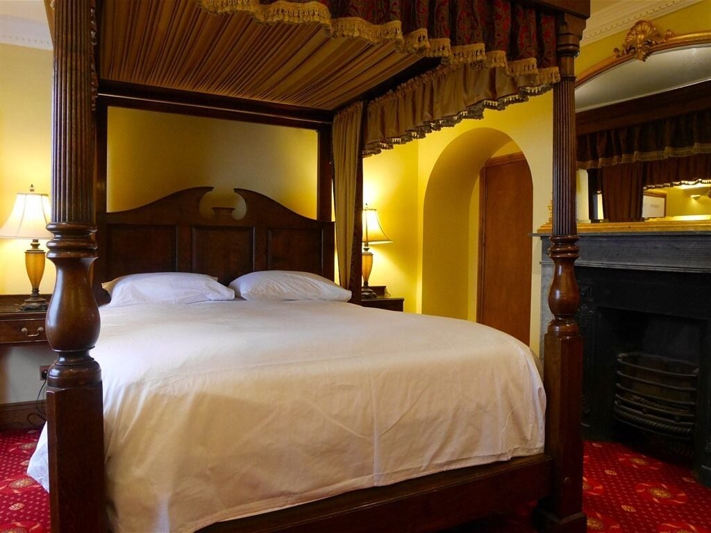 A four-poster bed with a wooden headboard, a red patterned carpet, and a fireplace with a mirror above it.