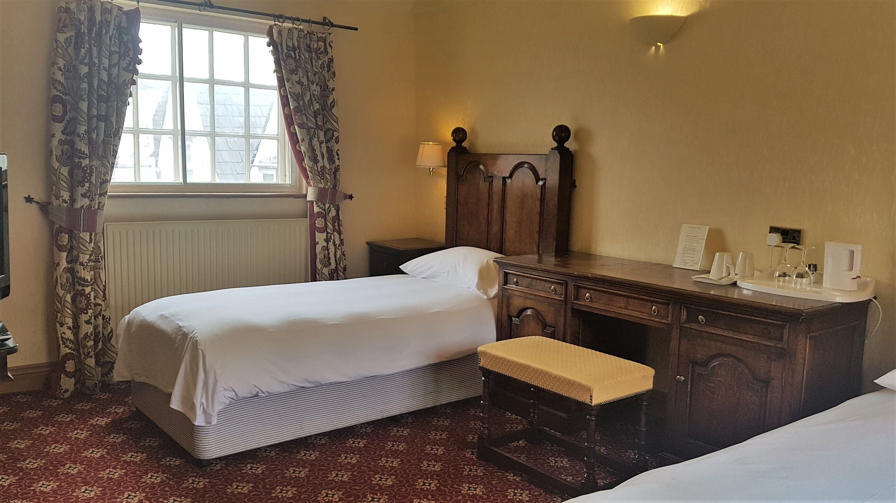 Une chambre d’hôtel avec un lit, une table de chevet en bois, un banc, une commode et une fenêtre avec des rideaux.