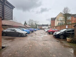 Um estacionamento com vários carros estacionados em filas, calçada de tijolos e edifícios ao fundo.