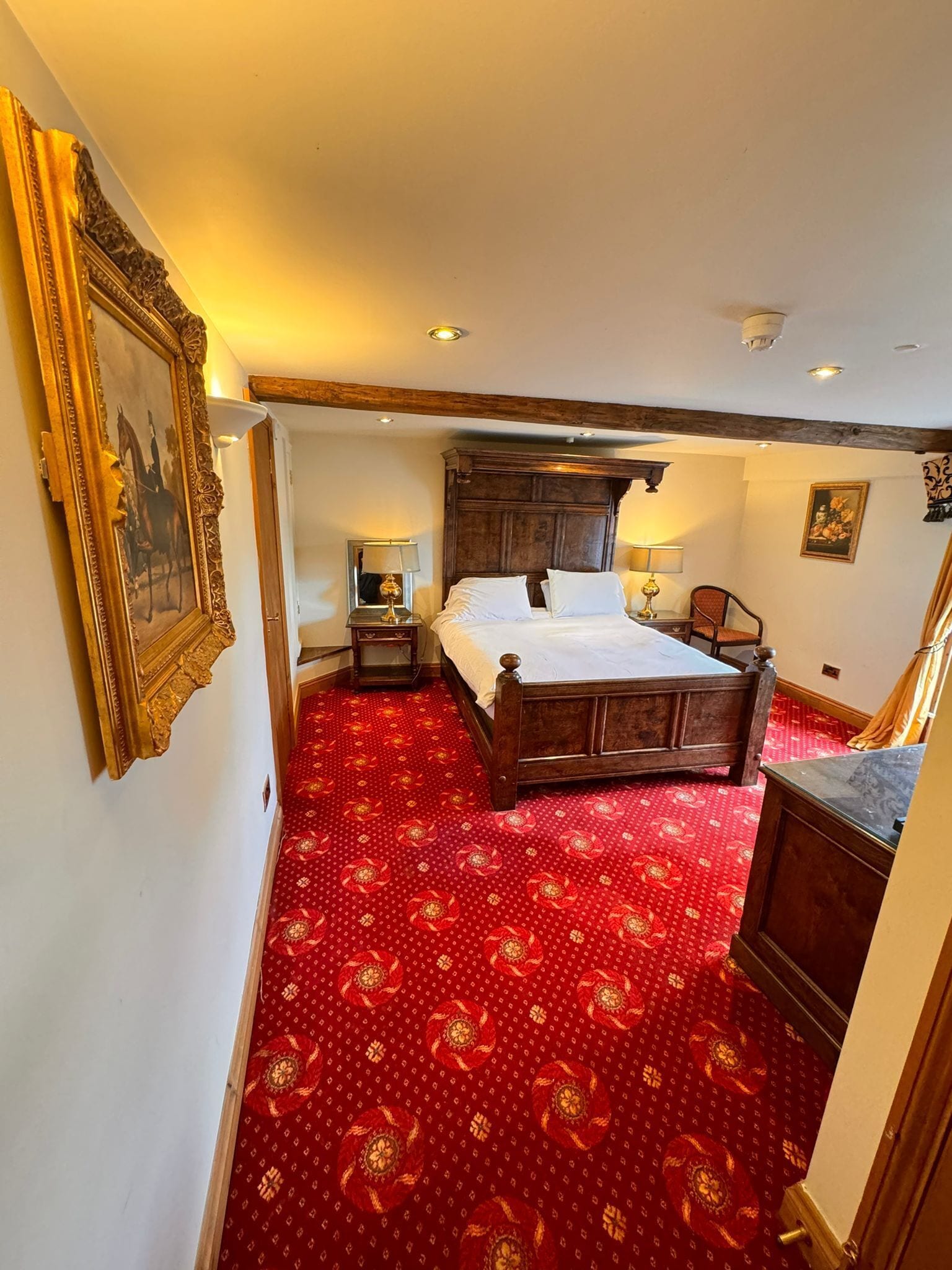 A bedroom with a large bed, a wooden headboard, a red patterned carpet, a nightstand with a lamp, and a framed picture on the wall.