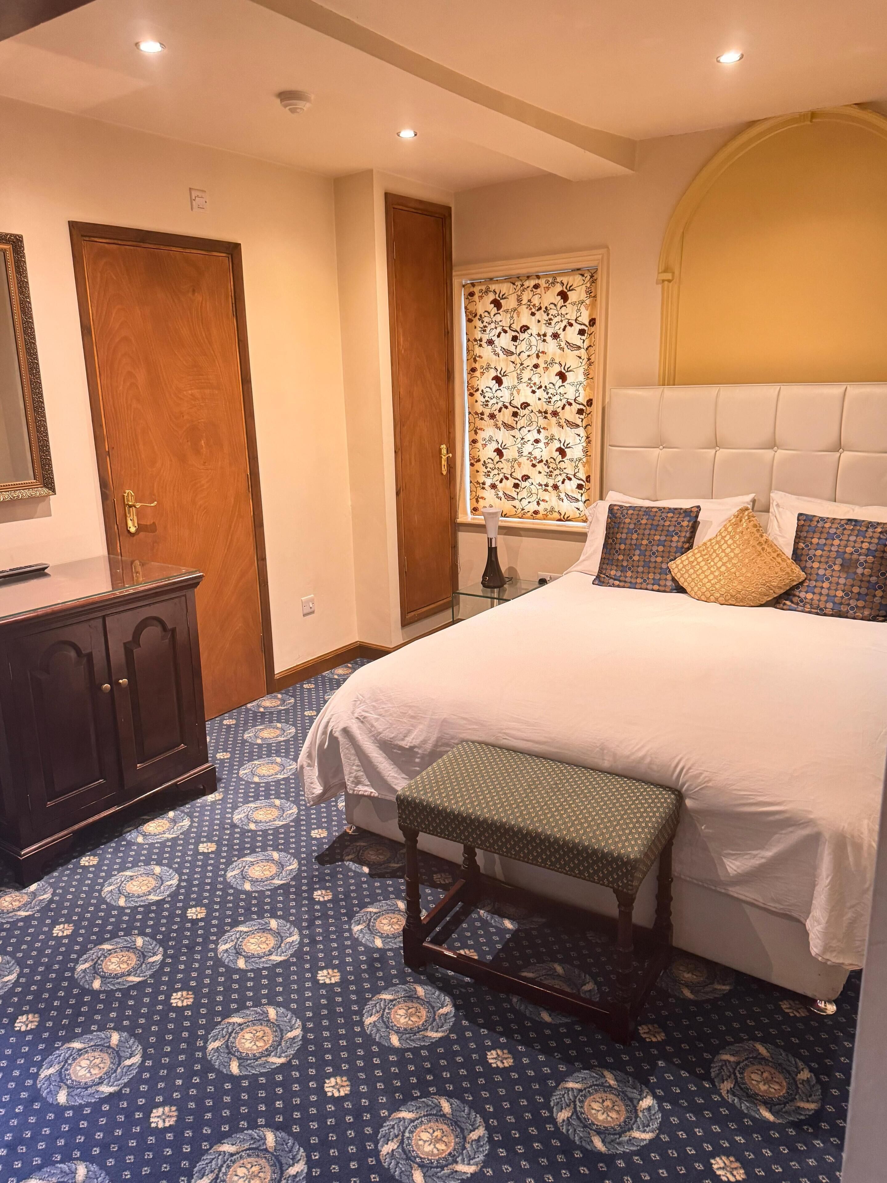 A bedroom with a large bed, a bench, a dresser, and a window with floral curtains.