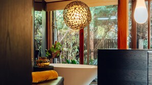 A modern bathroom with a freestanding bathtub, a hanging light fixture, and a view of greenery through large windows. - Sticky Rice Villas (Stirling)