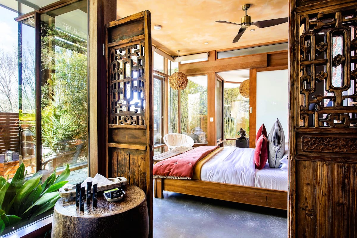 A spacious bedroom with a large bed, wooden furniture, and a view of greenery through large windows.