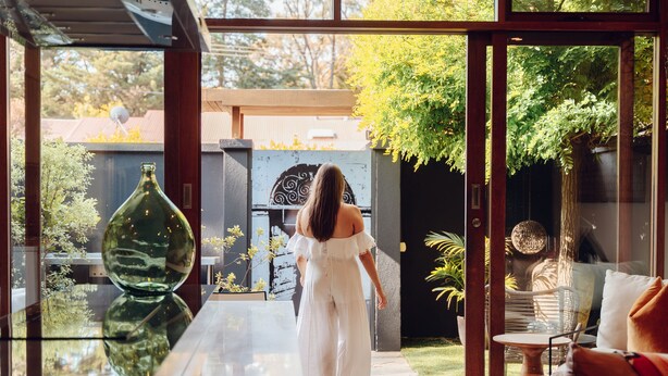 A woman in a white dress walking through a glass door into a garden area with a glass table and chairs. - Sticky Rice Villas (Stirling)