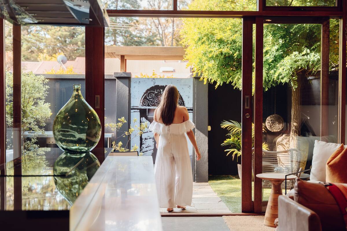 A woman in a white dress walking through a glass door into a garden area with a glass table and chairs.