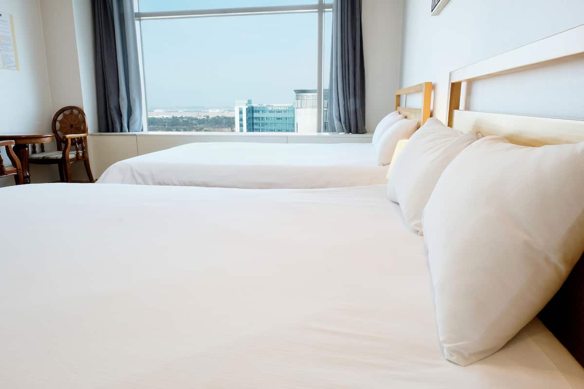 A hotel room with two beds, a wooden headboard, a view of the cityscape, and a small table with a chair.