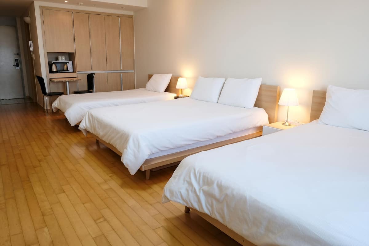A hotel room with three single beds, a wooden floor, a desk with a chair, and a wardrobe.