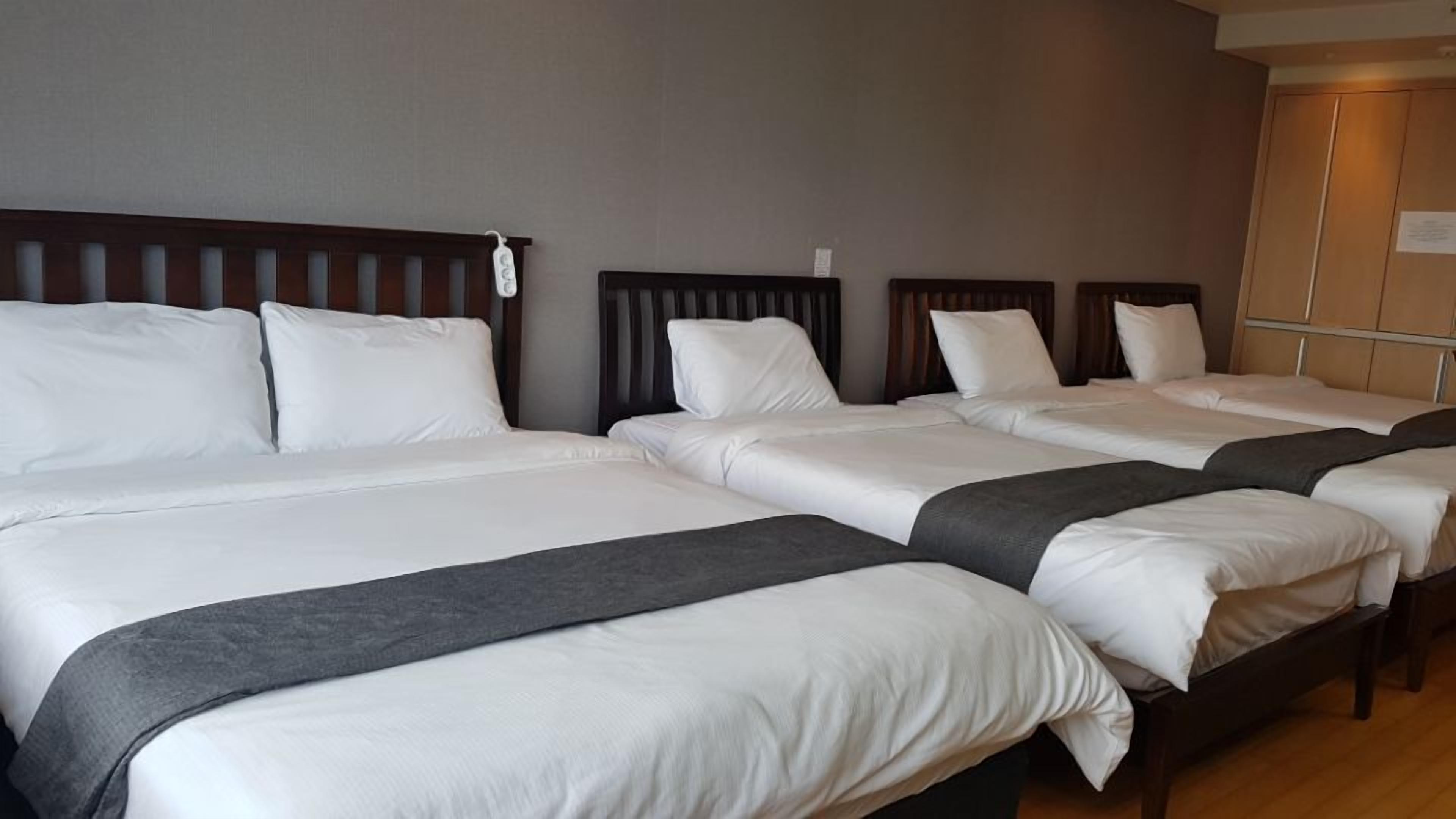 A hotel room with four single beds aligned in a row, each with white bedding and a dark headboard.