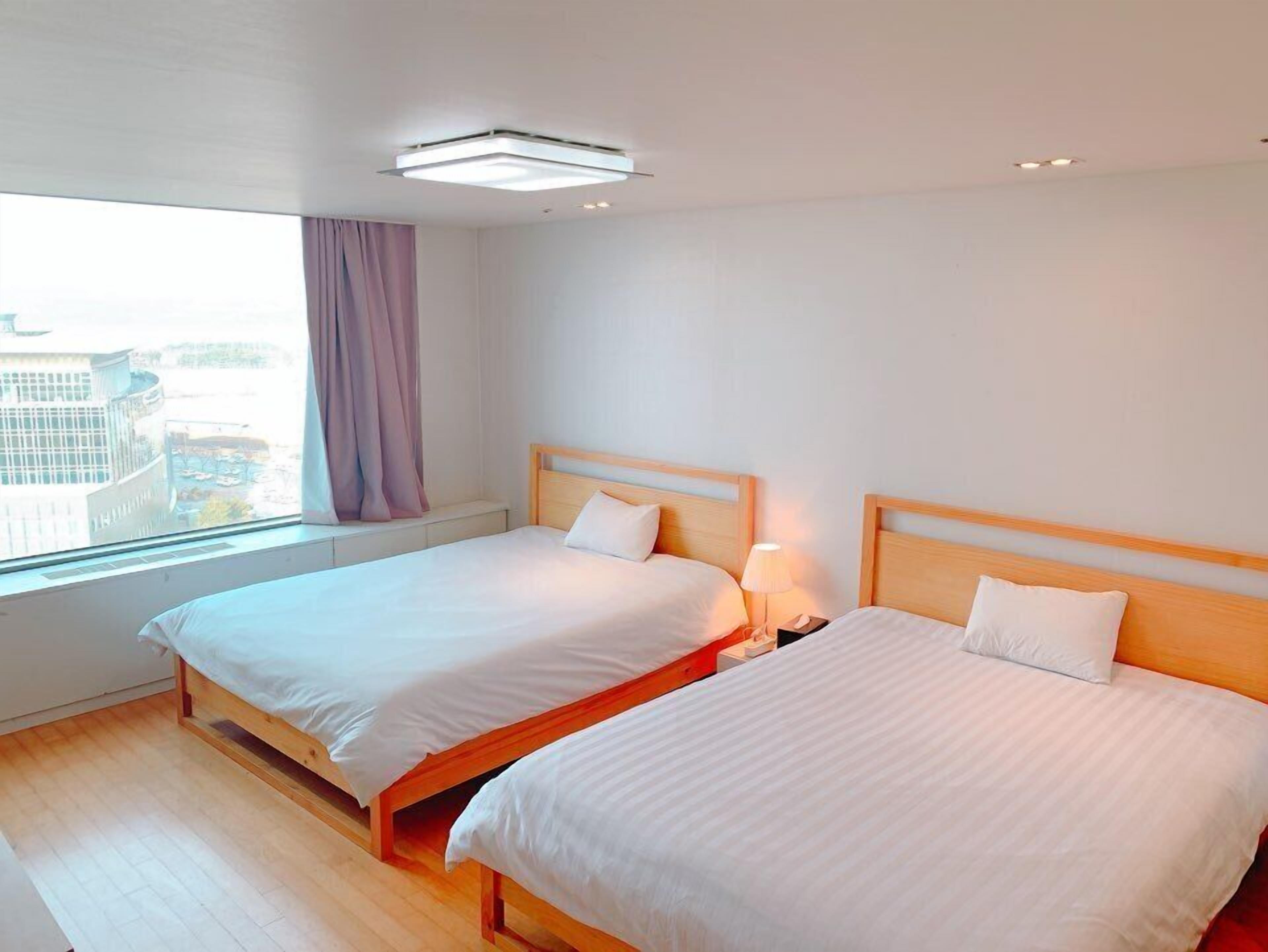 A hotel room with two beds, a window with a view of buildings, a ceiling light, and a nightstand with a lamp.