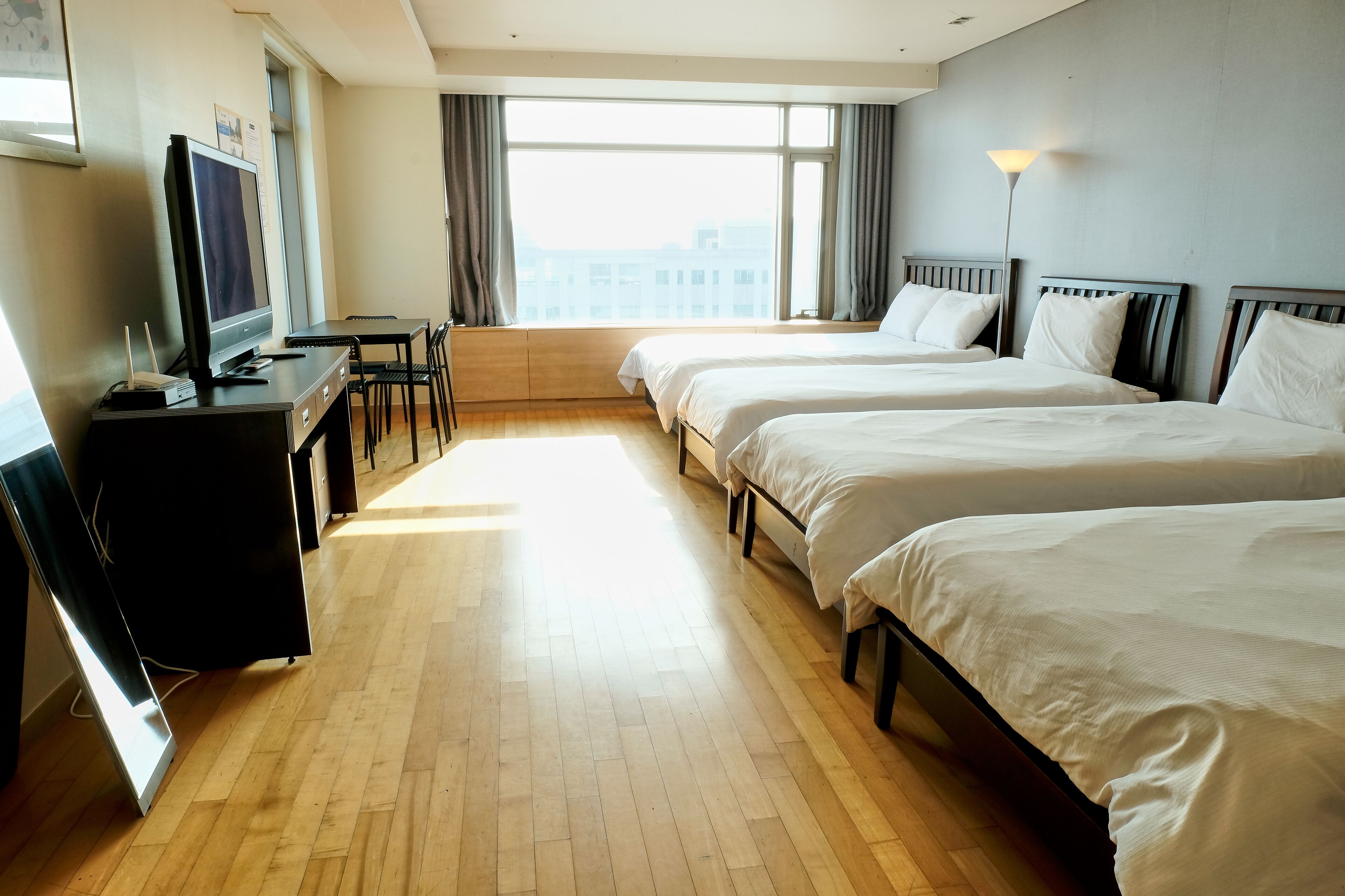 A hotel room with two beds, a desk with a computer, a TV, and a large window with curtains.