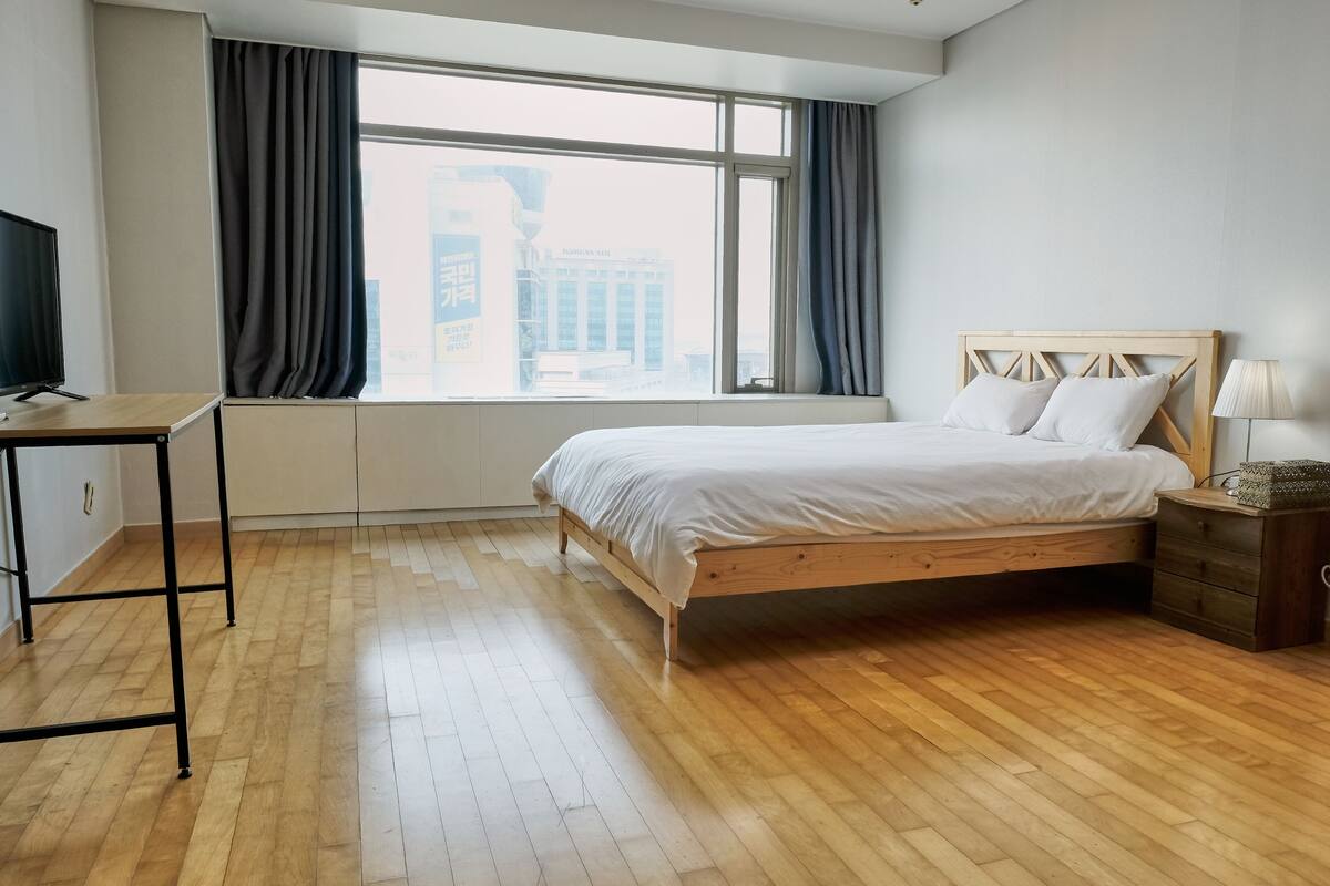 A modern hotel room with a wooden floor, a bed with white bedding, a desk with a TV, and a large window with curtains.