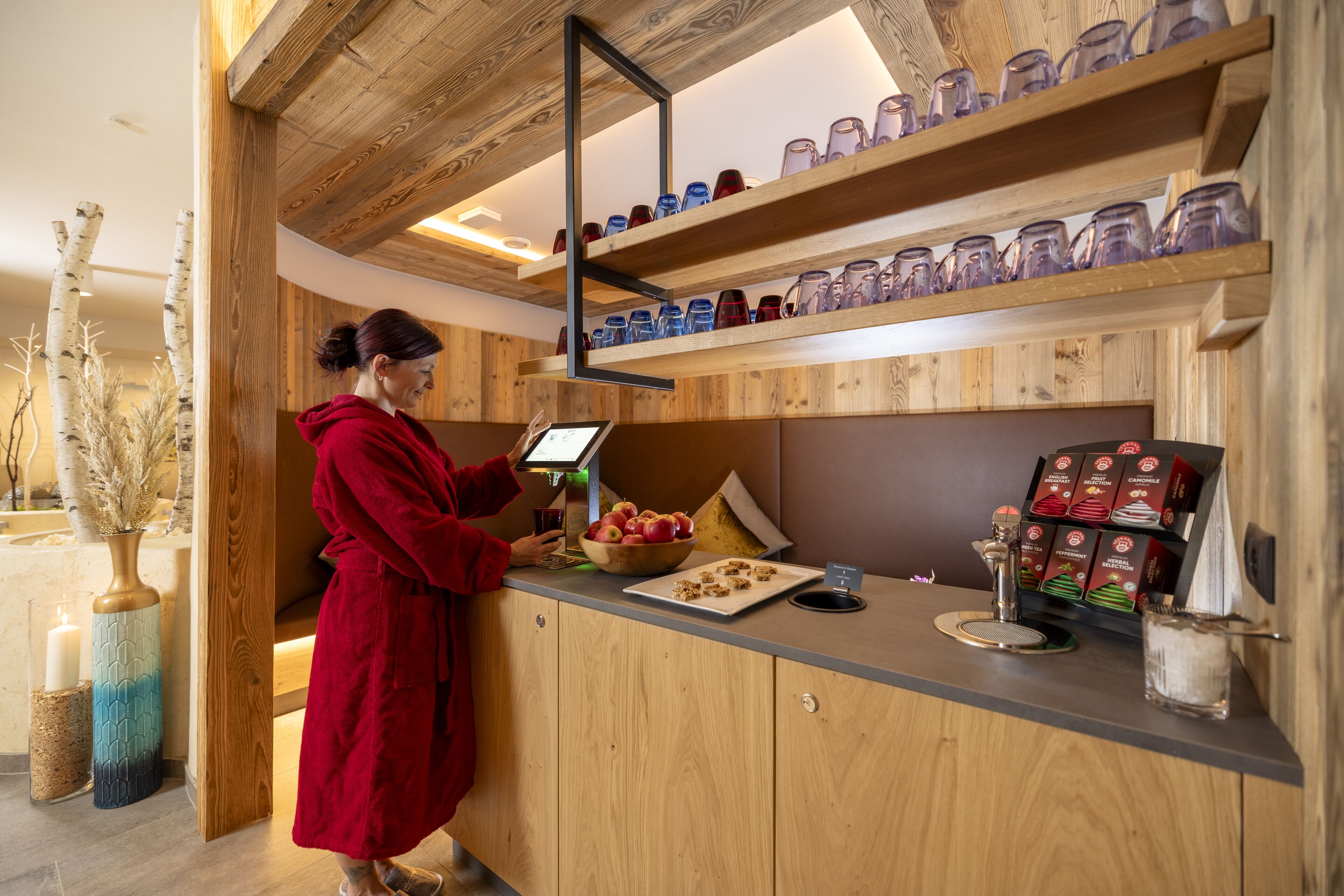 Interior de madera con una persona vestida con una bata roja en un mostrador, junto a un bol de fruta, una tableta y una máquina de café.