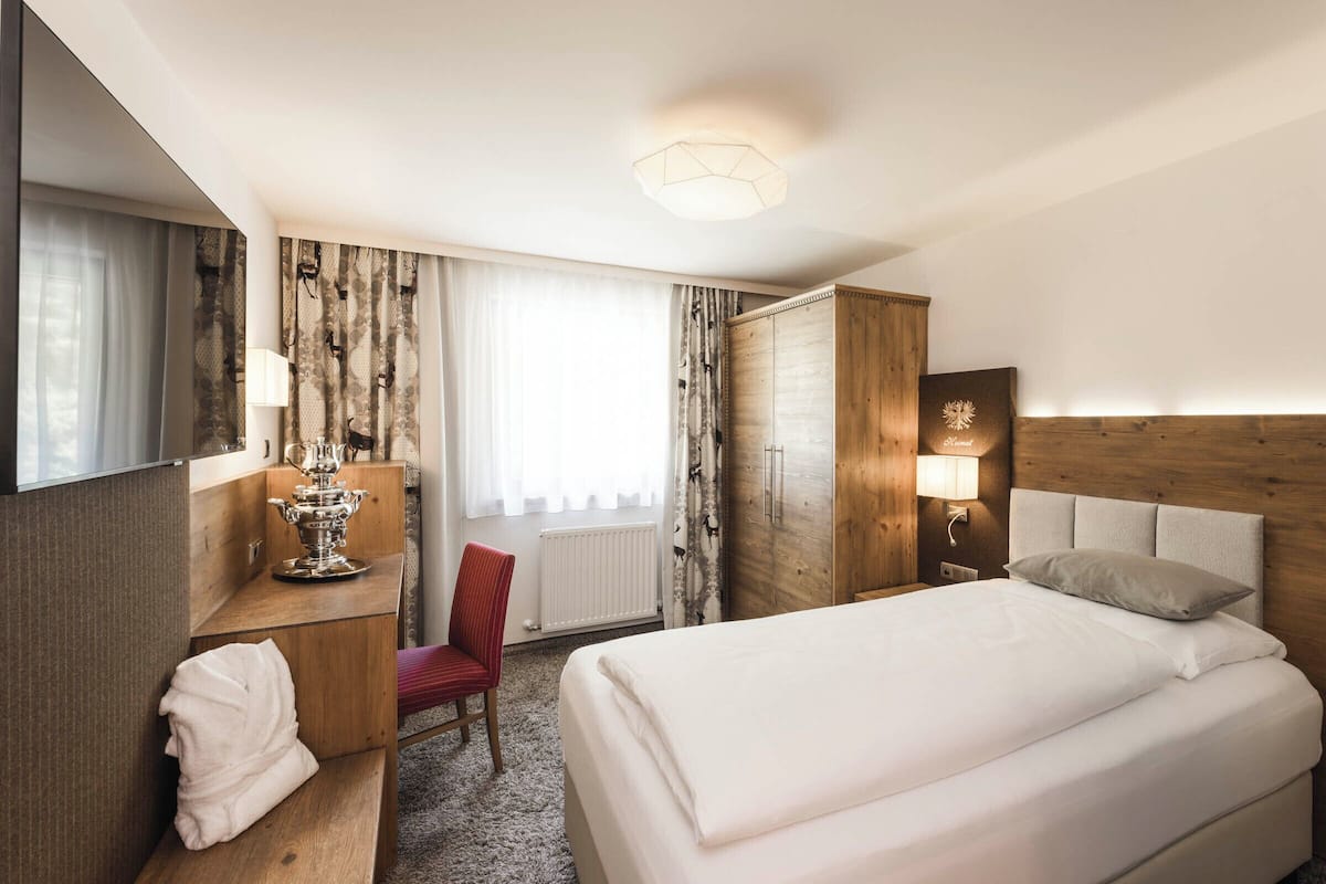 A hotel room with a large bed, a wooden wardrobe, a desk with a lamp, a chair, and a window with curtains.