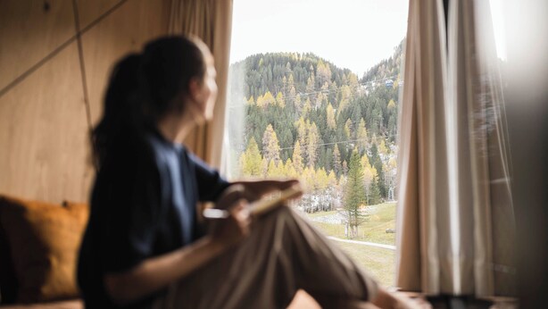 A person sitting on a bed in a room with a large window, looking out at a forested hillside. -  VALARA Alpine Art Hotel (Tux)