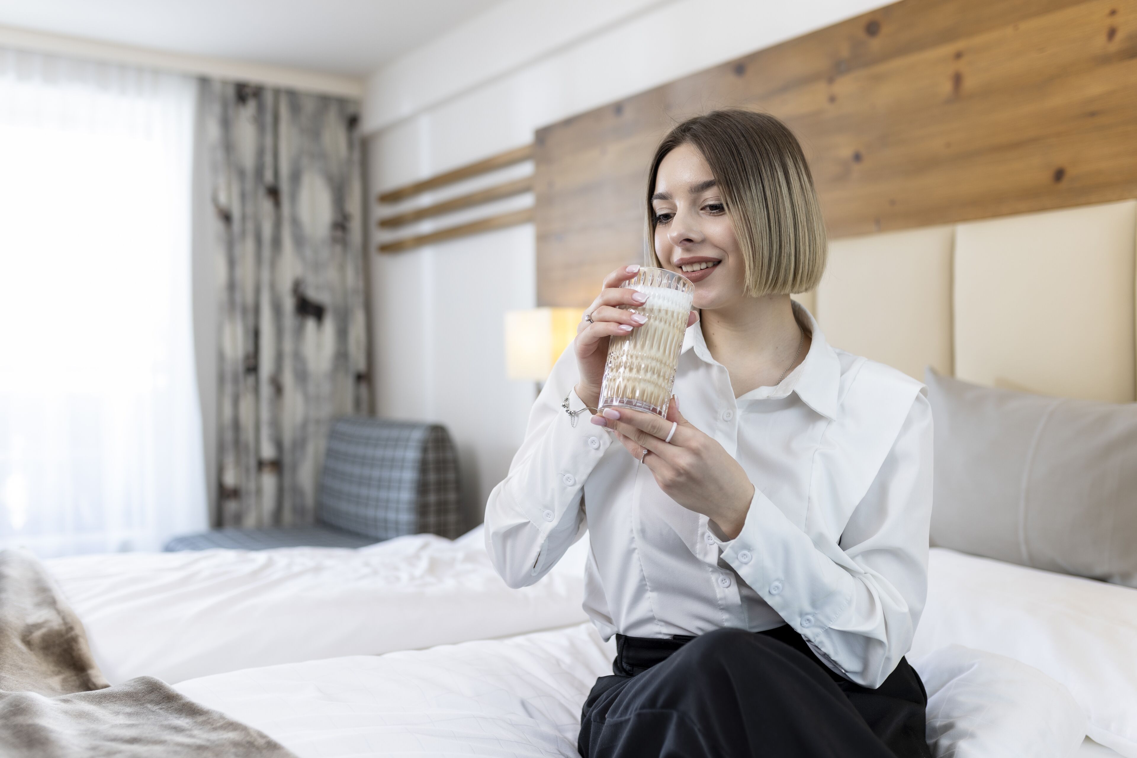 A person sitting on a bed in a hotel room with a wooden headboard, holding a glass of water.