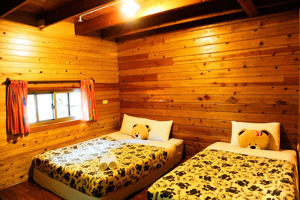 A cabin with two beds, each with a teddy bear pillow, wooden walls, and a window with curtains.