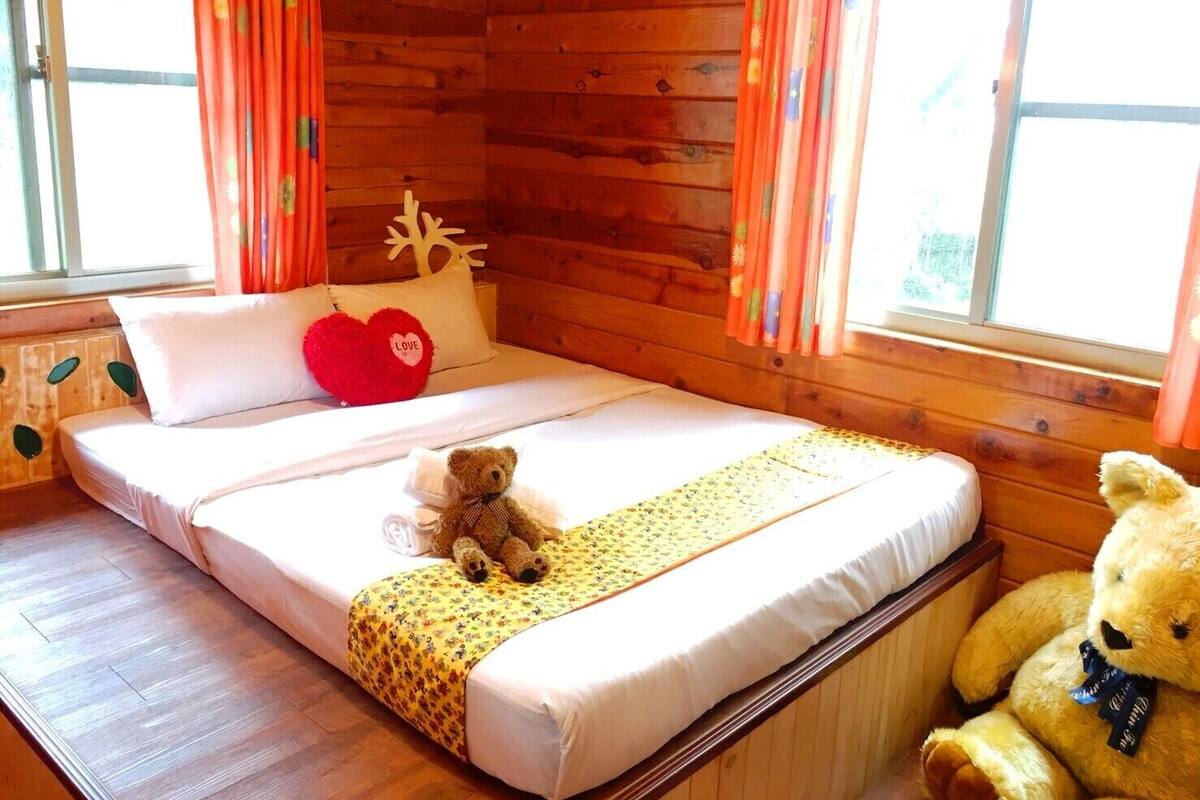A wooden cabin room with a bed, a teddy bear, and a large teddy bear on the floor.