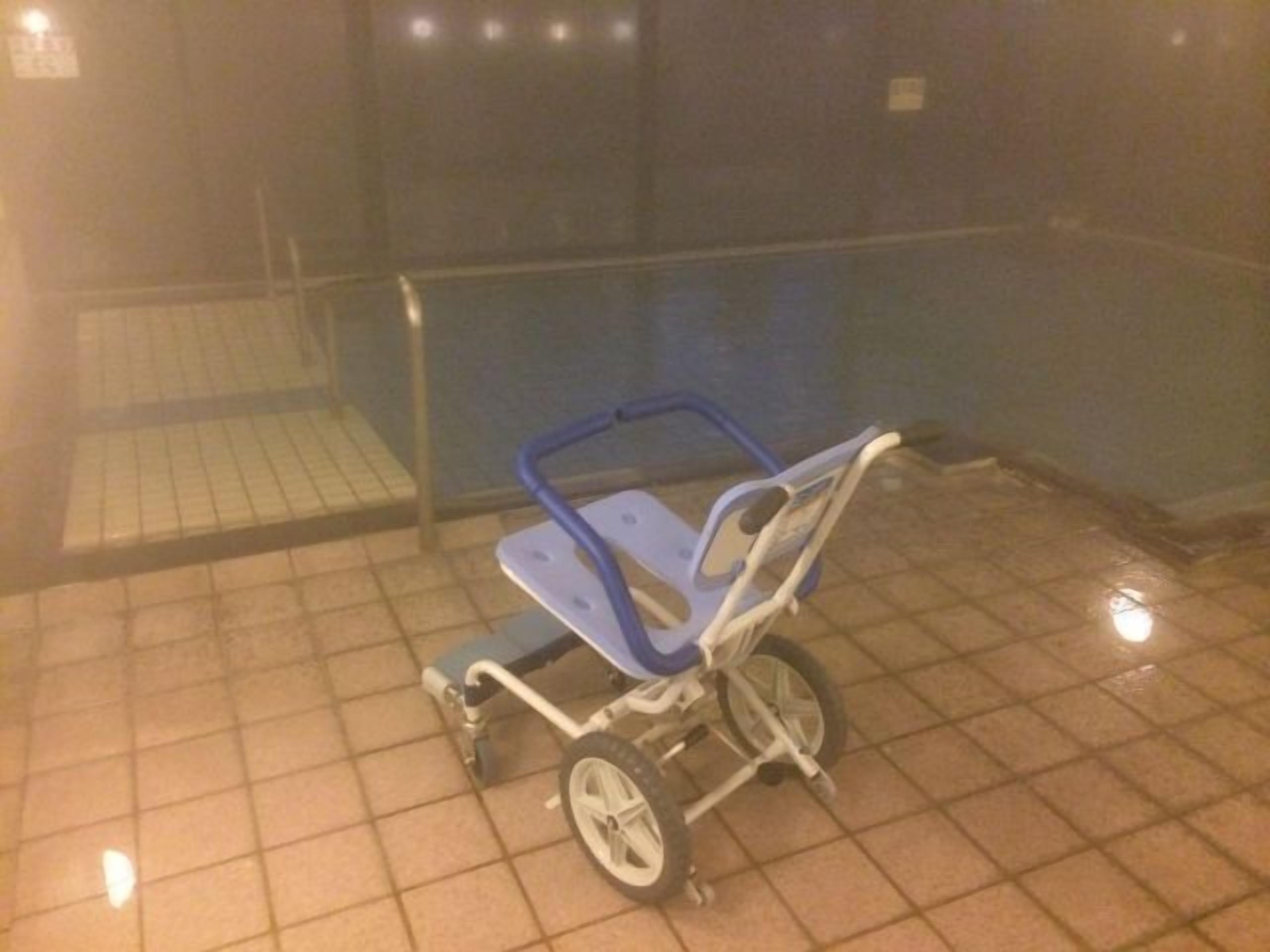 Una sedia a rotelle in una stanza scarsamente illuminata, con pavimento piastrellato e una porta in vetro.
