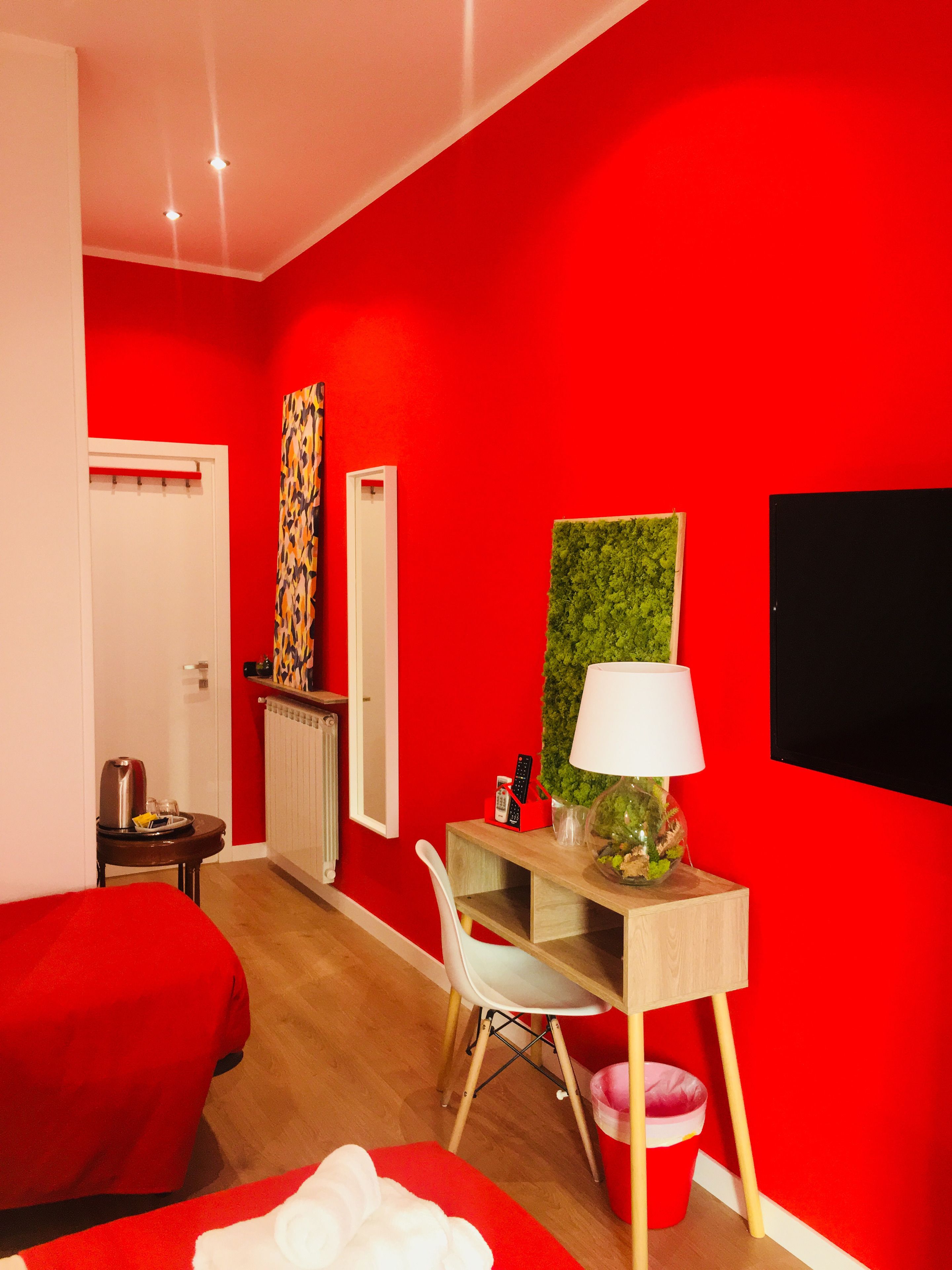 A room with red walls, a bed, a desk with a lamp, a chair, a radiator, and a television.