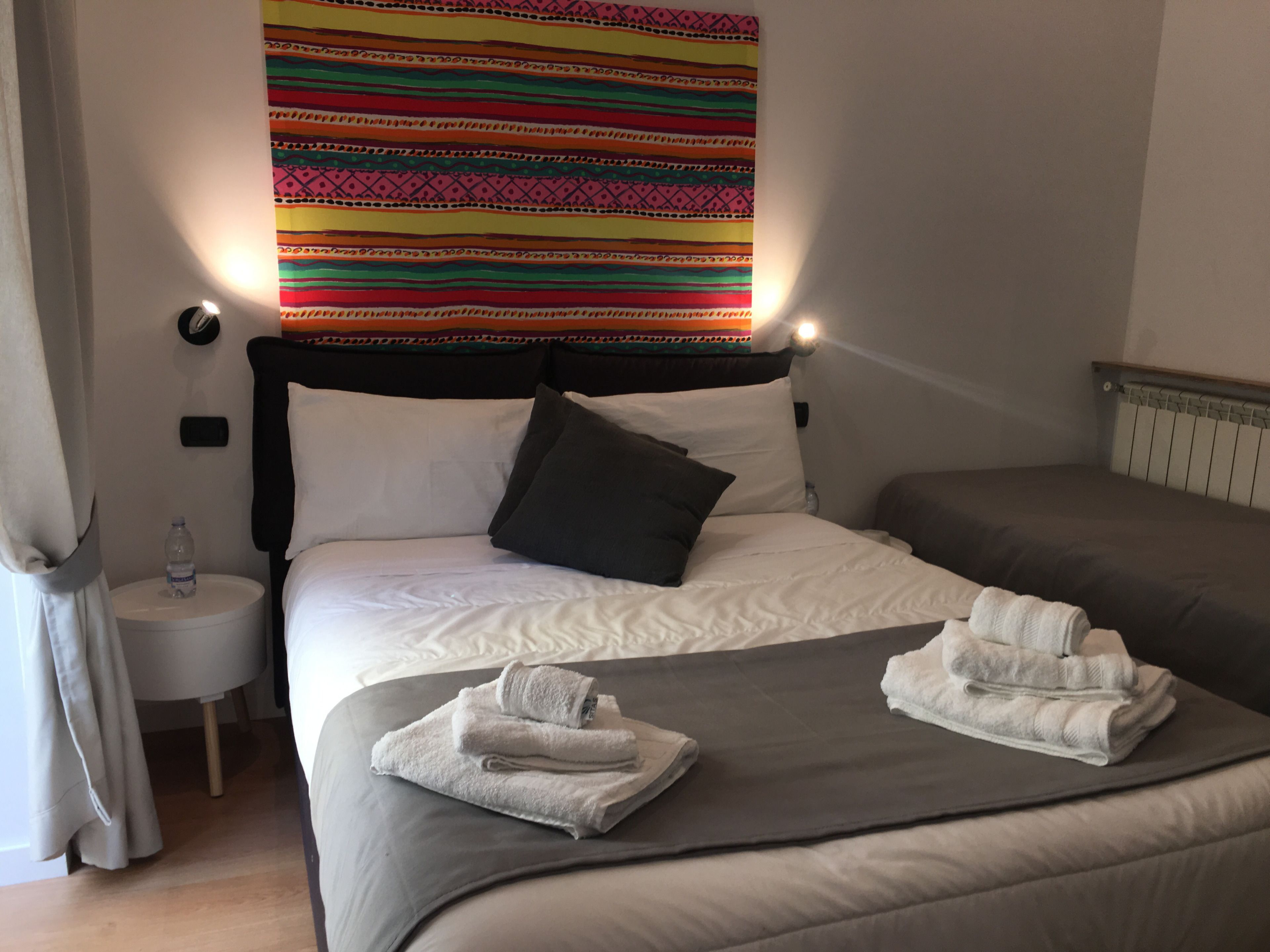 A hotel room with a bed, two pillows, a headboard, a bedside table with a water bottle, and a colorful tapestry on the wall.