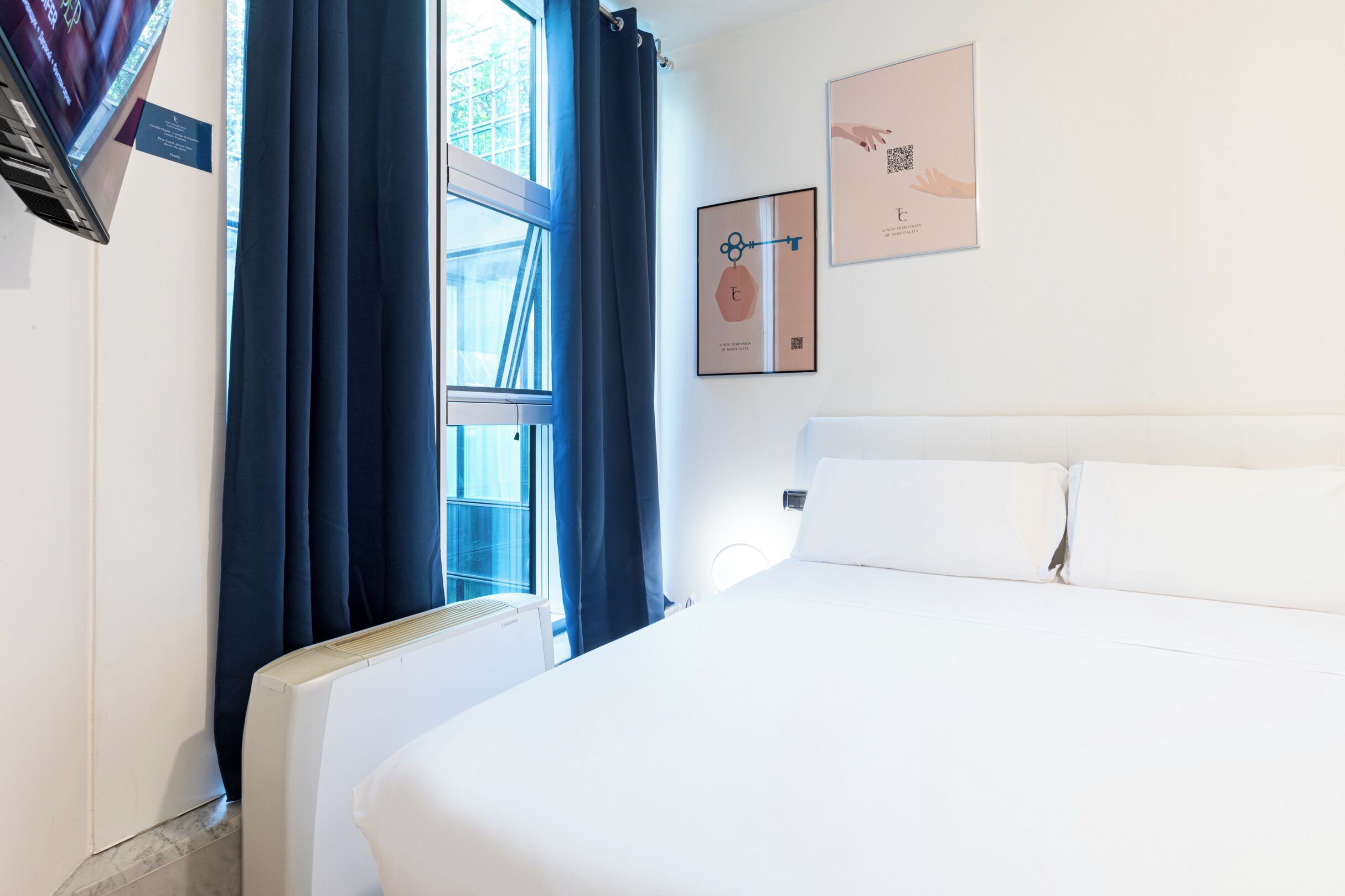 Une chambre d’hôtel avec un lit, une fenêtre avec des rideaux, un radiateur et des œuvres d’art murales.