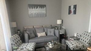A living room with a grey sofa, patterned armchairs, a coffee table, and a side table with a lamp.