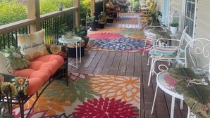 A covered porch with outdoor seating, a colorful rug, and various plants.