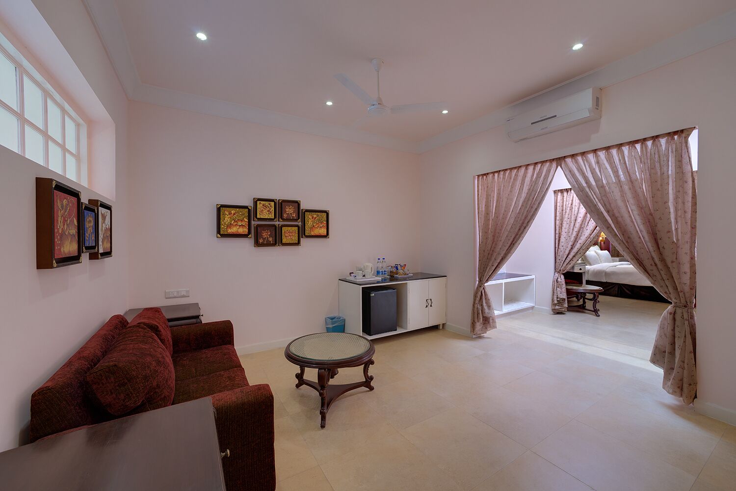 A spacious living room with a red sofa, a small round table, and a TV on a cabinet. There are framed pictures on the wall and a bed in the background.