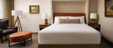 A hotel room with a large bed, a chair, a small ottoman, two nightstands with lamps, and a framed picture on the wall.