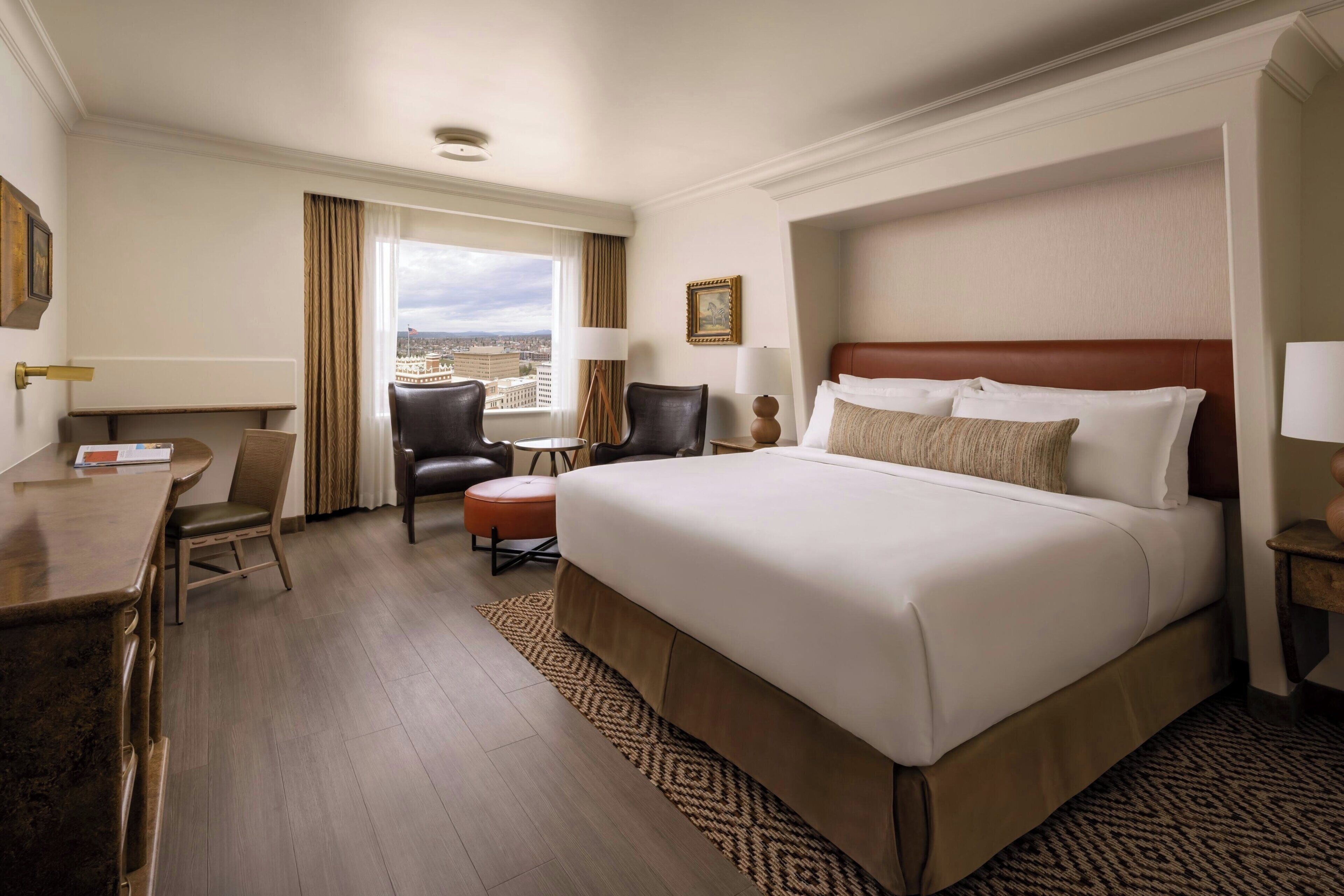 A hotel room with a large bed, a desk, two chairs, a small table, and a view of the city.