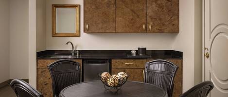 A dining area with a round table, four chairs, a bowl of decorative items, a black countertop, a sink, and wooden cabinets.