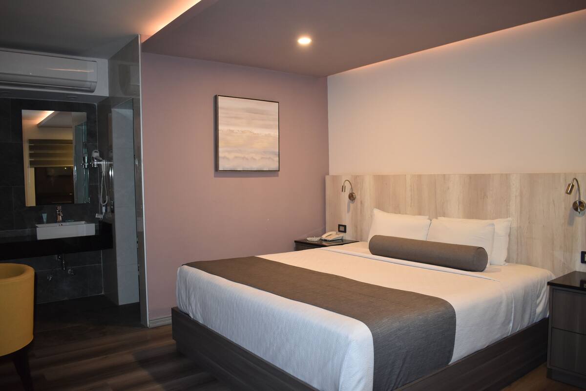 A modern hotel room with a large bed, a bedside table, a nightstand, a mirror, and a bathroom with a sink and shower.