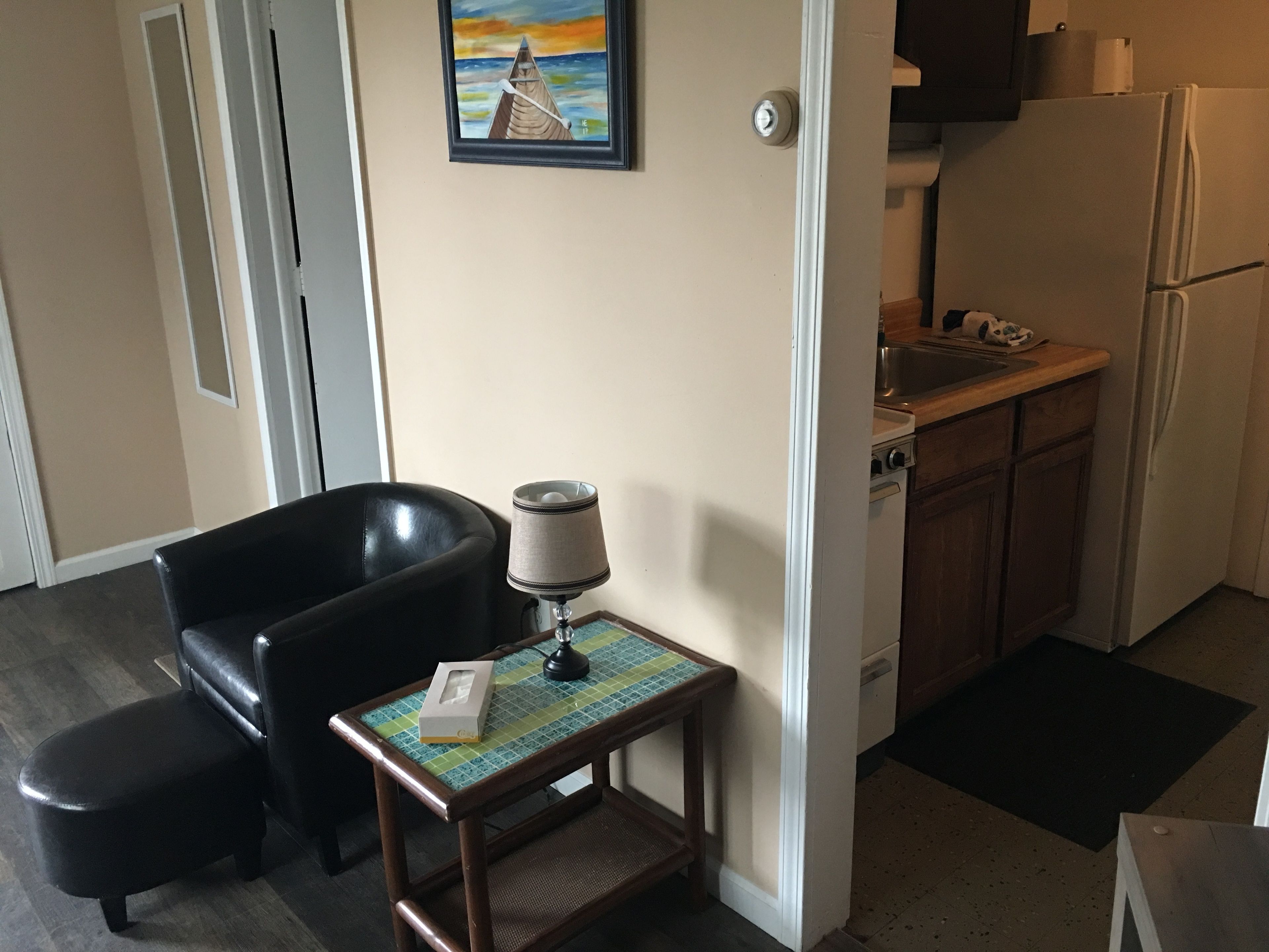 A compact living space with a kitchenette, a black leather chair, a small table with a lamp, and a painting on the wall.