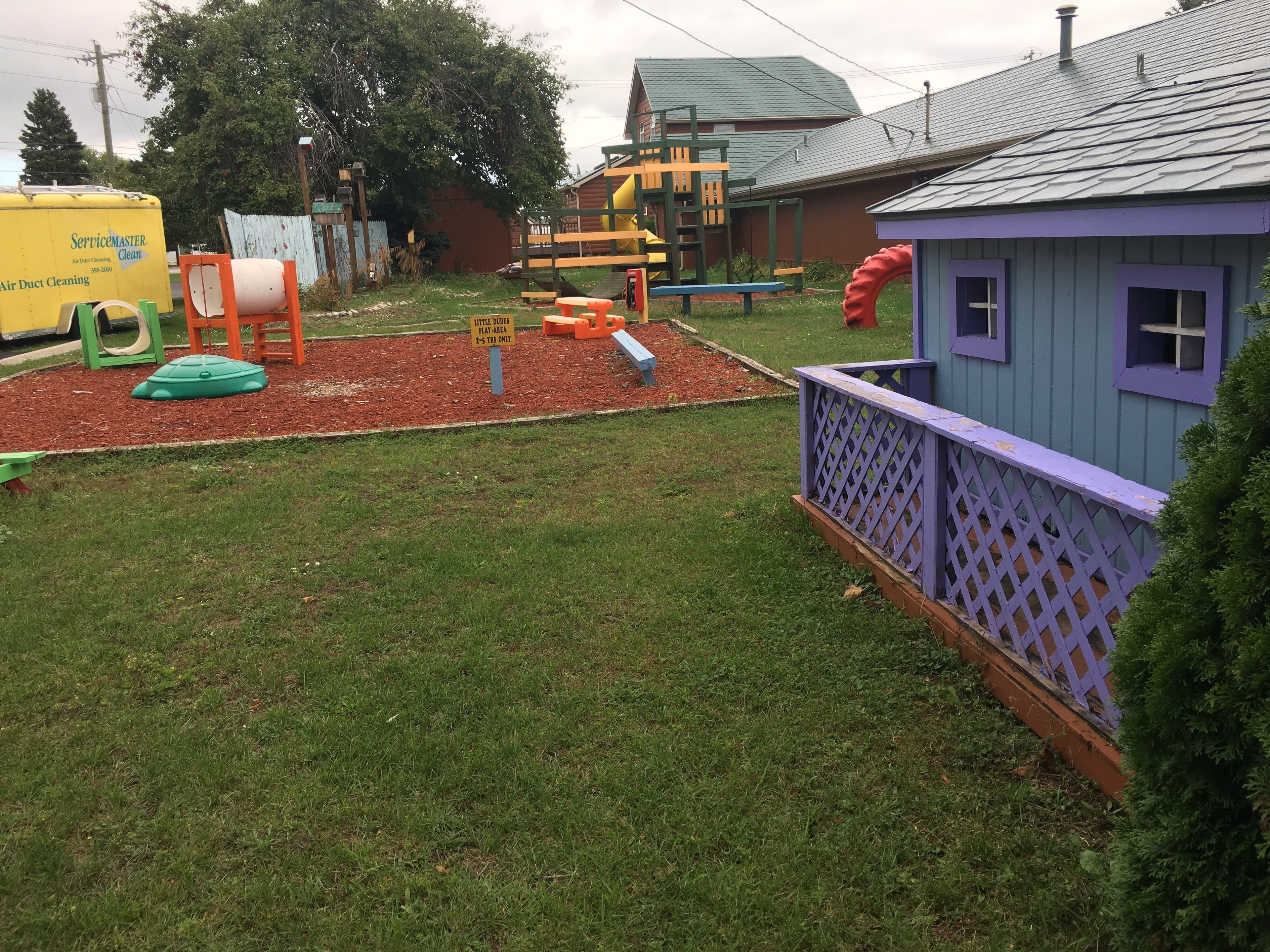 A playground with various play structures, a small building with purple trim, and a yellow service truck in the background.