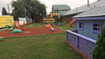 A playground with various play structures, a small building with purple trim, and a yellow service truck in the background.