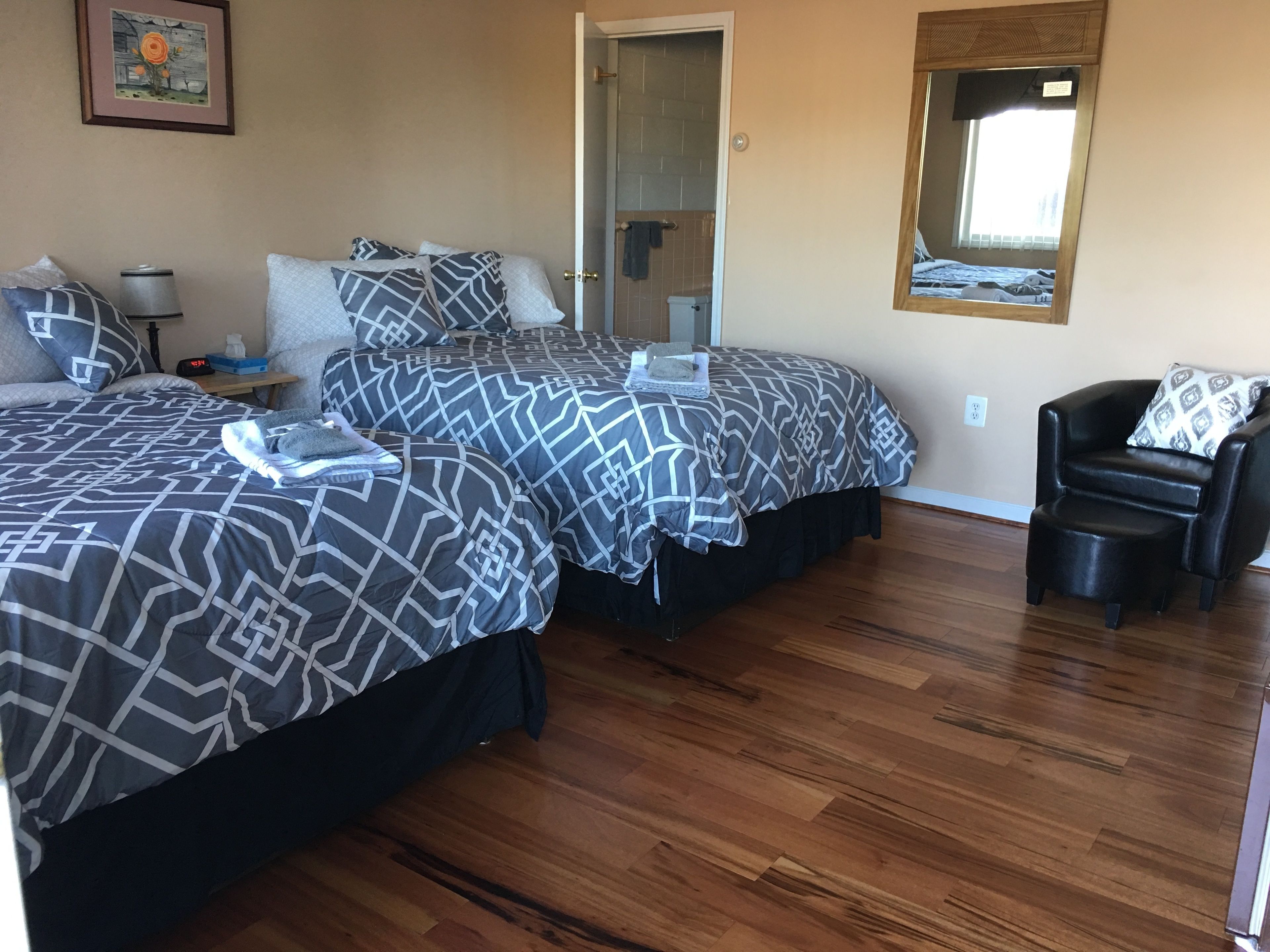 Habitación de hotel con dos camas, piso de madera, una silla, una mesita de noche, una lámpara y un cuadro en la pared.