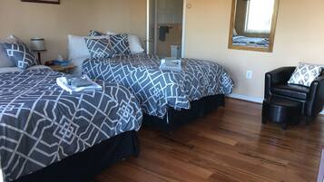 Habitación de hotel con dos camas, piso de madera, una silla, una mesita de noche, una lámpara y un cuadro en la pared.