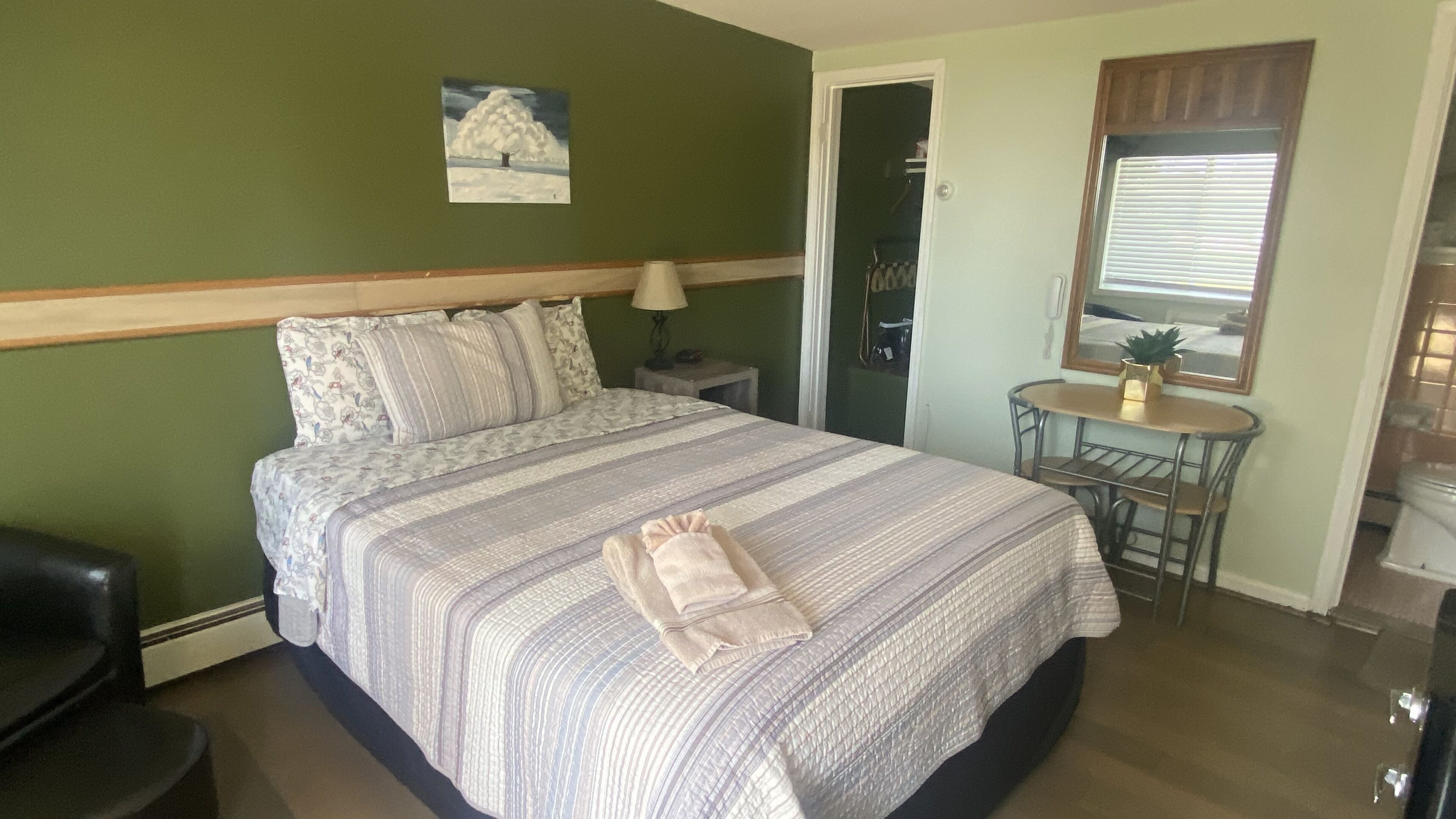 Una cama bien tendida con cubrecama y almohadas a rayas, una mesita con una silla, un cuadro en la pared y una puerta que da al baño.