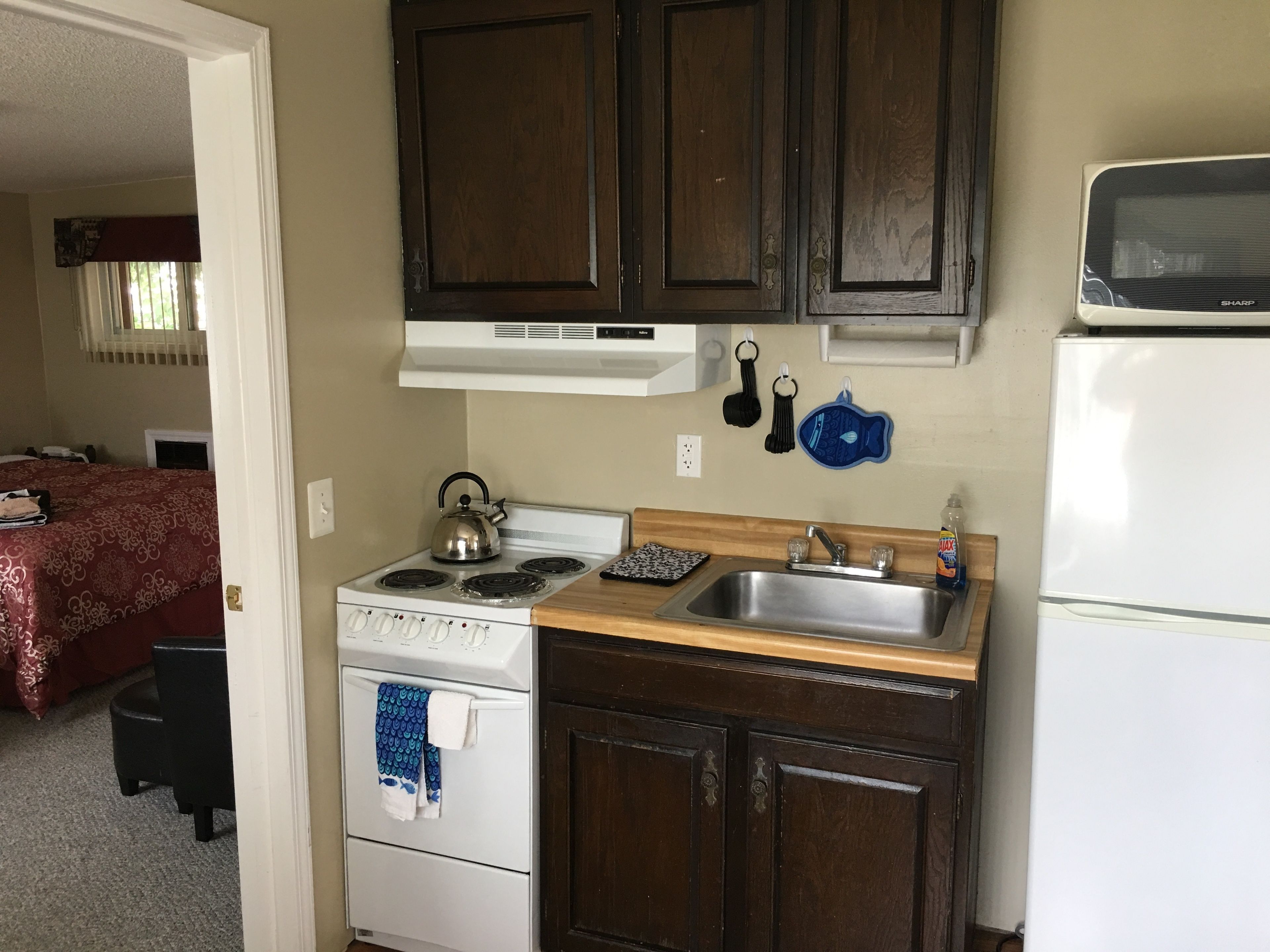 Una cocina compacta con estufa, pileta y microondas. Hay muebles altos de madera sobre la encimera y una cama con colcha roja en la habitación contigua.