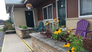 A building with a brick facade, a black door, and a window with blinds. There are purple Adirondack chairs and a planter with flowers on the porch. - Big Bear Lodge (Alpena)