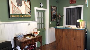 A reception area with a wooden desk, a clock, framed artwork, a sign prohibiting pets, and a window with curtains. - School House Inn (North Conway)