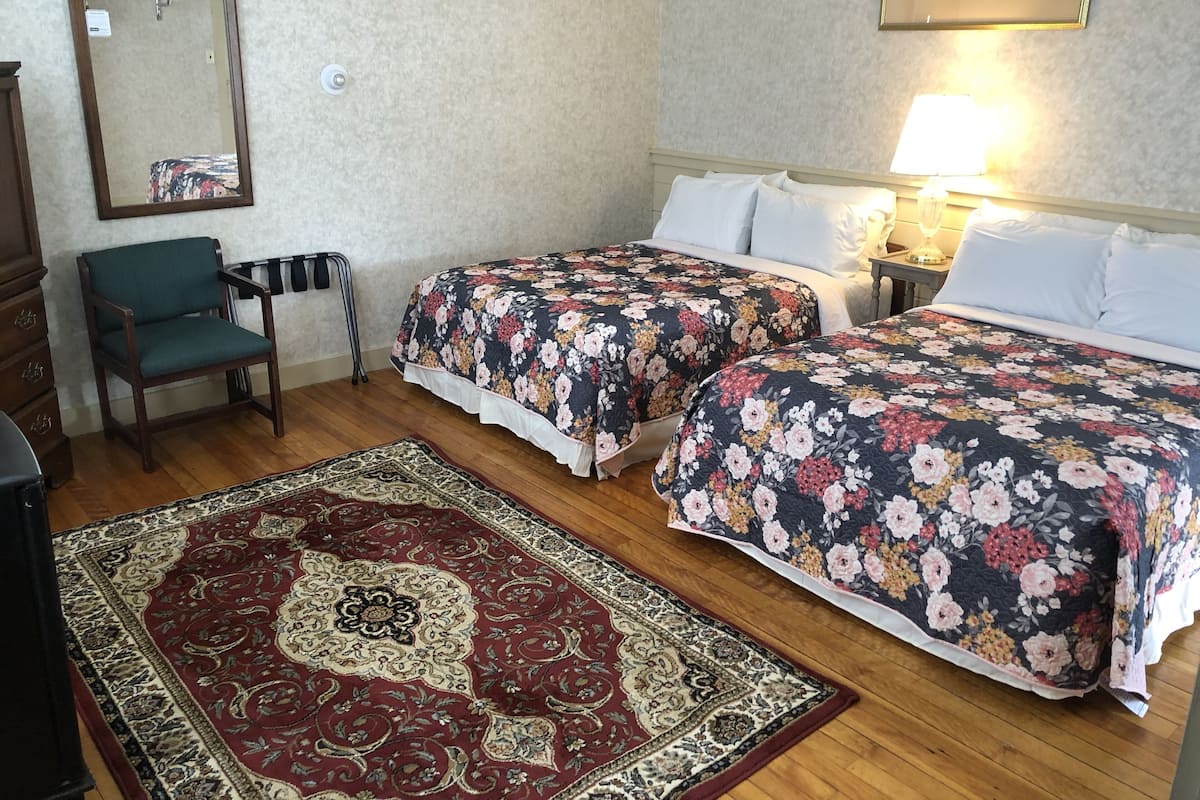 A hotel room with two beds, a chair, a mirror, a lamp, and a patterned rug.