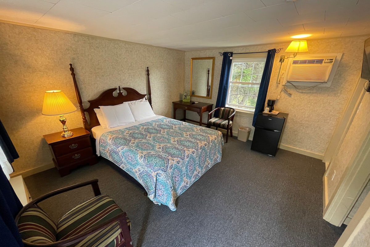 A neatly arranged hotel room with a bed, a chair, a desk, a lamp, and a window with curtains.