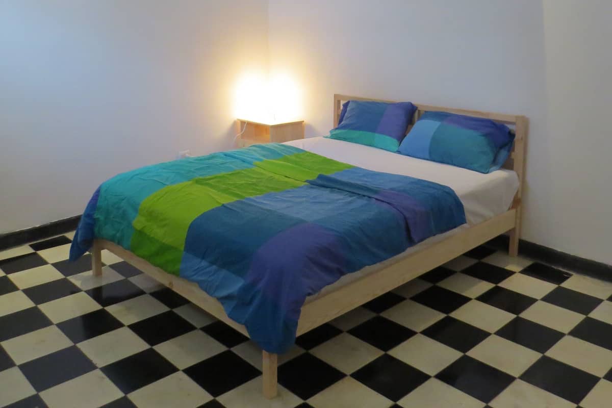 Una cama individual con estructura de madera, suelo de cuadros y una mesita de noche con lámpara.
