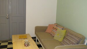 Ein Zimmer mit einem Schachbrettmuster auf dem Boden, einem Holztisch mit einem Glasbehälter, einem beige farbenen Sofa mit gestreiften Kissen und einer grauen Tür.