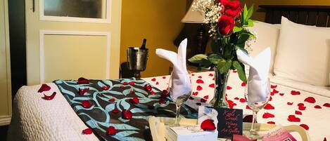 A neatly made bed with rose petals, a vase of flowers, and a tray with a book and a glass of wine.