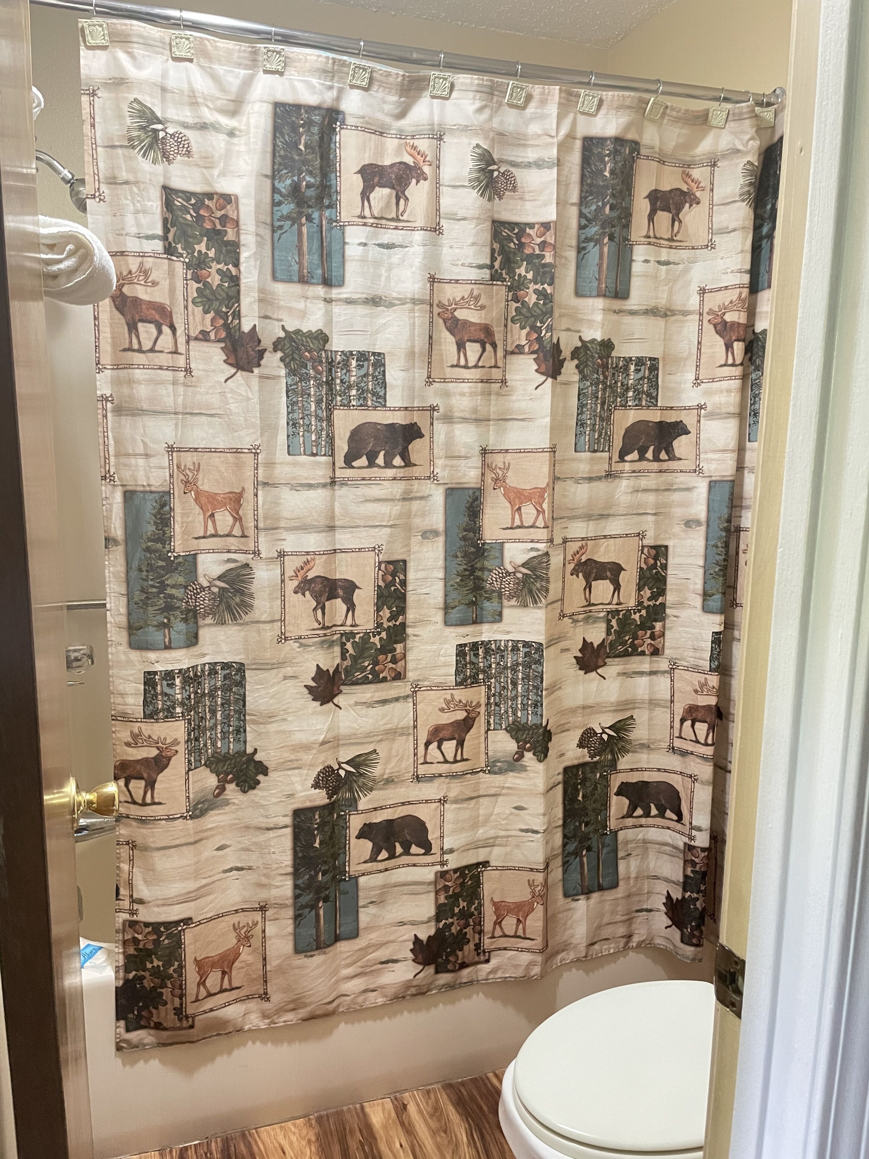 A bathroom with a shower curtain featuring a forest and wildlife print, a toilet, and a wooden floor.